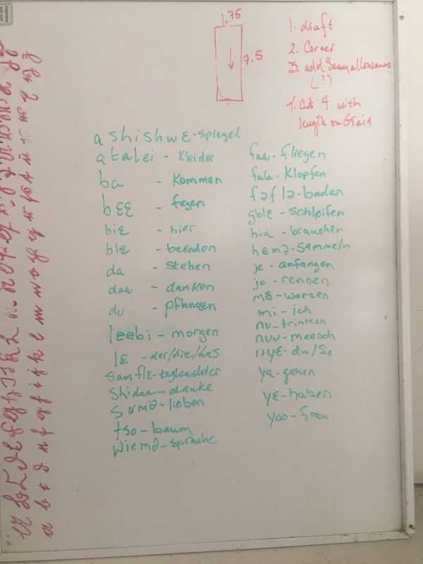 Vocabulary list in Ga and German on whiteboard next to zipper facing instructions at Tchad sewing studio workroom in Chicago